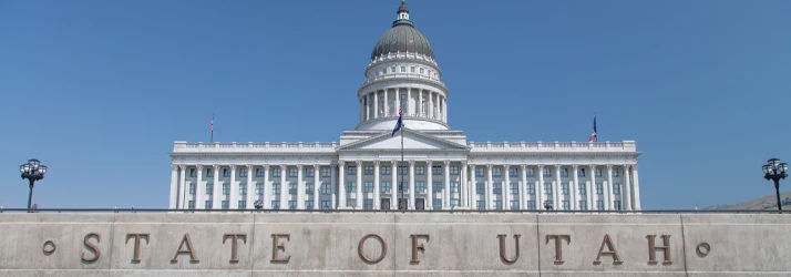 White domed government building with columns behind a stone wall displaying “STATE OF UTAH.”