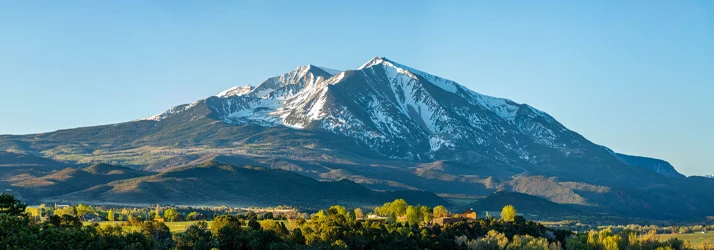 Snow-capped mountain peak rising above rolling hills and trees under a clear blue sky