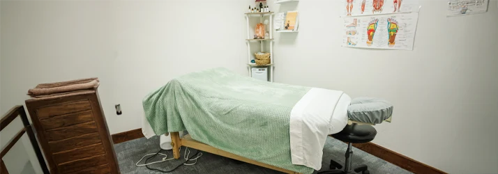 Treatment room with an examination table, stool, shelving unit, and anatomical charts on the wall.