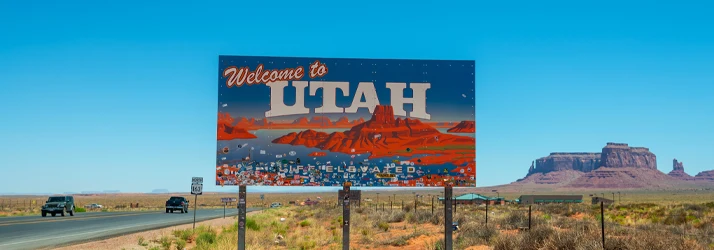 Roadside billboard reading “Welcome to Utah” with desert landscape and mesas in the background.